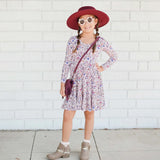 Young girl in a floral dress, red hat, and sunglasses standing against a white brick wall.