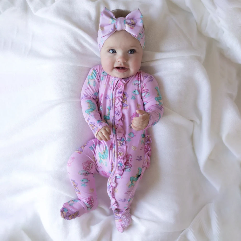 A baby lying on white bedding, wearing a pink footed sleeper with a mermaid print and a matching pink bow headband.