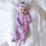 A baby lying on white bedding, wearing a pink footed sleeper with a mermaid print and a matching pink bow headband.