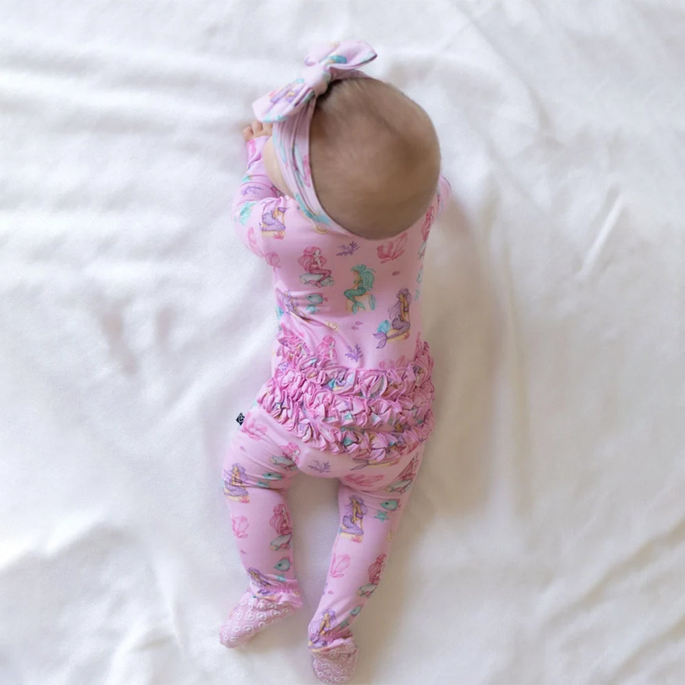 A baby lying on a white fabric surface, wearing a pink footed bodysuit with a mermaid print, ruffled waist, and matching pink bow headband.