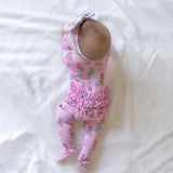 A baby lying on a white fabric surface, wearing a pink footed bodysuit with a mermaid print, ruffled waist, and matching pink bow headband.