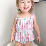 A smiling toddler girl wearing a pink ruffle-strap romper with a mermaid and sea life print, standing indoors against a light gray wall.
