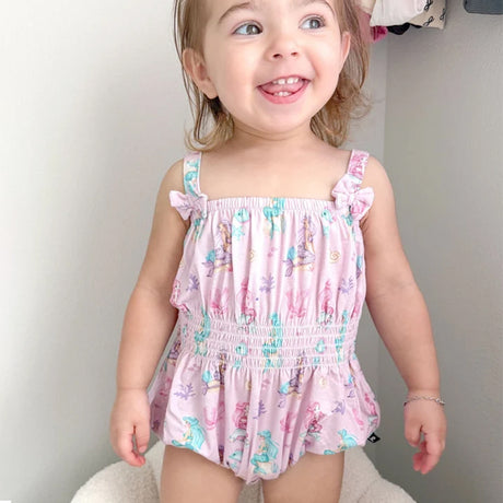 A smiling toddler girl wearing a pink ruffle-strap romper with a mermaid and sea life print, standing indoors against a light gray wall.