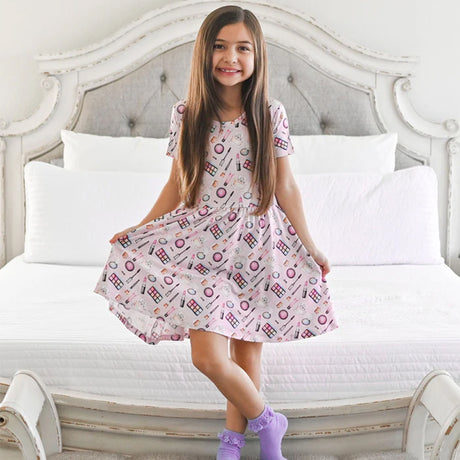 Young girl in a pink makeup-themed dress sitting on a bed, applying makeup.