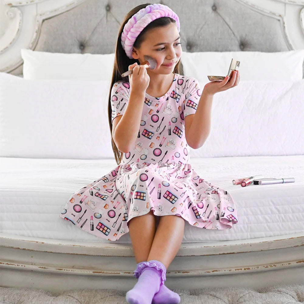 Young girl in a pink makeup-themed dress sitting on a bed, applying makeup.