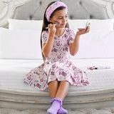 Young girl in a pink makeup-themed dress sitting on a bed, applying makeup.