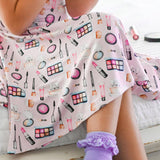 Person wearing a pink outfit with makeup-themed pattern sitting on a bed.