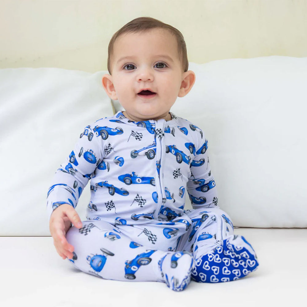 Baby wearing a white onesie with blue car patterns sitting on a white couch.
