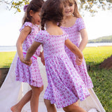 Three young girls in matching pink floral dresses standing outdoors near a body of water.