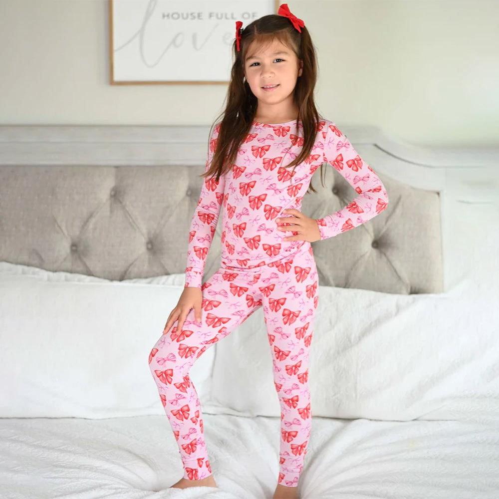 Young girl wearing pink bow patterned pajamas sitting on a bed.