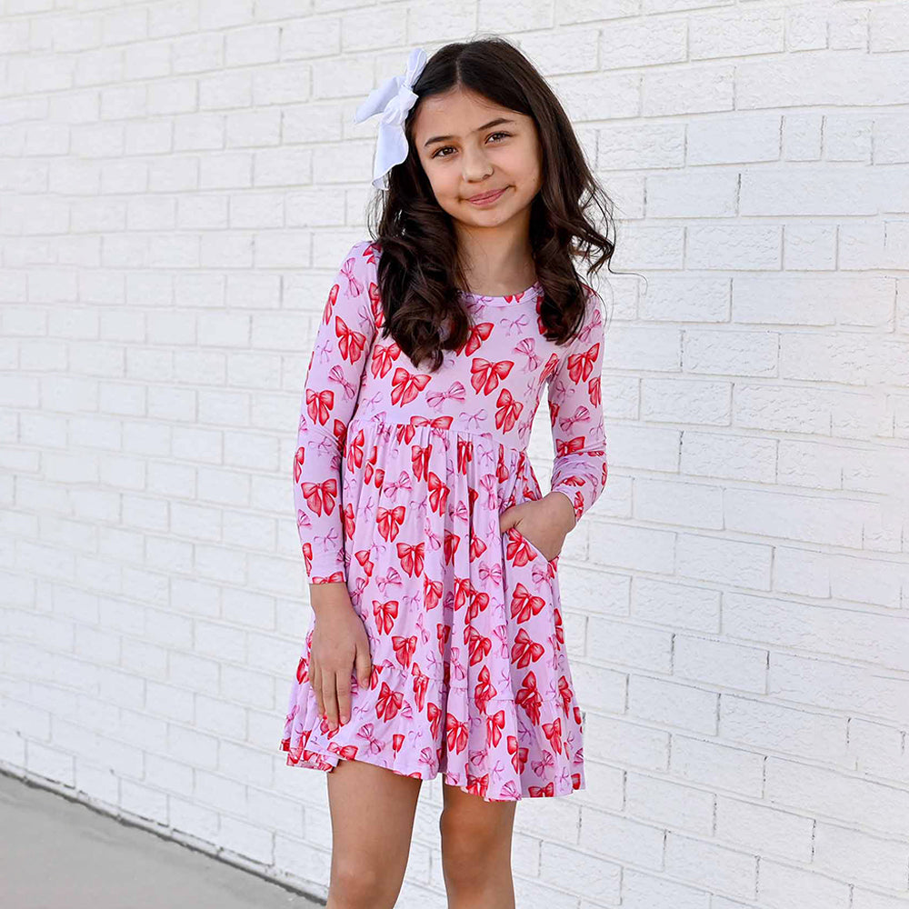 Young girl wearing a pink dress with red patterns against a white brick wall.