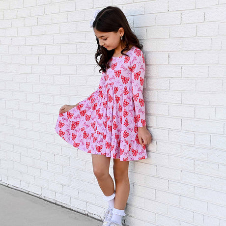Young girl wearing a pink dress with red patterns against a white brick wall.