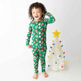 Child wearing a green Christmas-themed onesie standing next to a small decorated tree on a white background