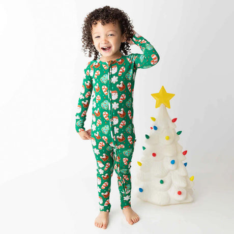 Child wearing a green Christmas-themed onesie standing next to a small decorated tree on a white background