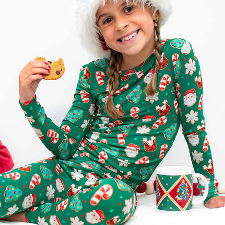 Child wearing a green Christmas-themed onesie holding a cookie and a mug.