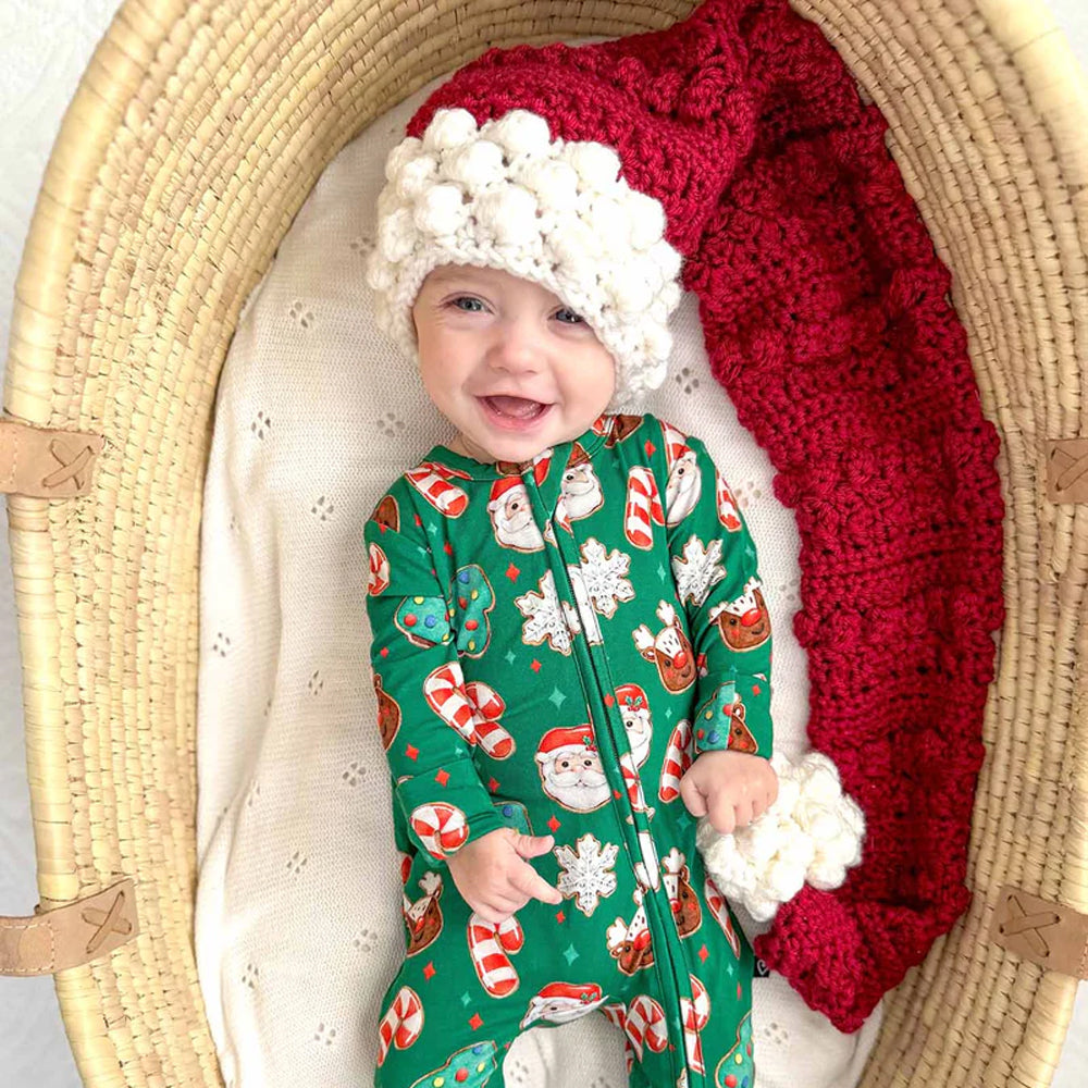Baby in a festive onesie with a green pattern, red blanket, and white hat, lying in a moses basket.
