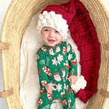 Baby in a festive onesie with a green pattern, red blanket, and white hat, lying in a moses basket.