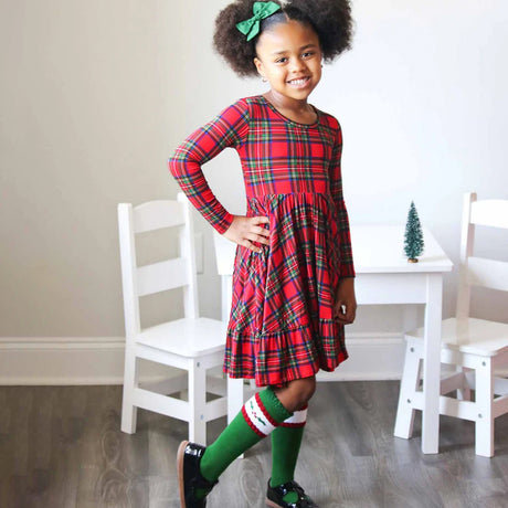 Young girl in a red plaid dress with green bow standing in a room with white chairs and a small table.