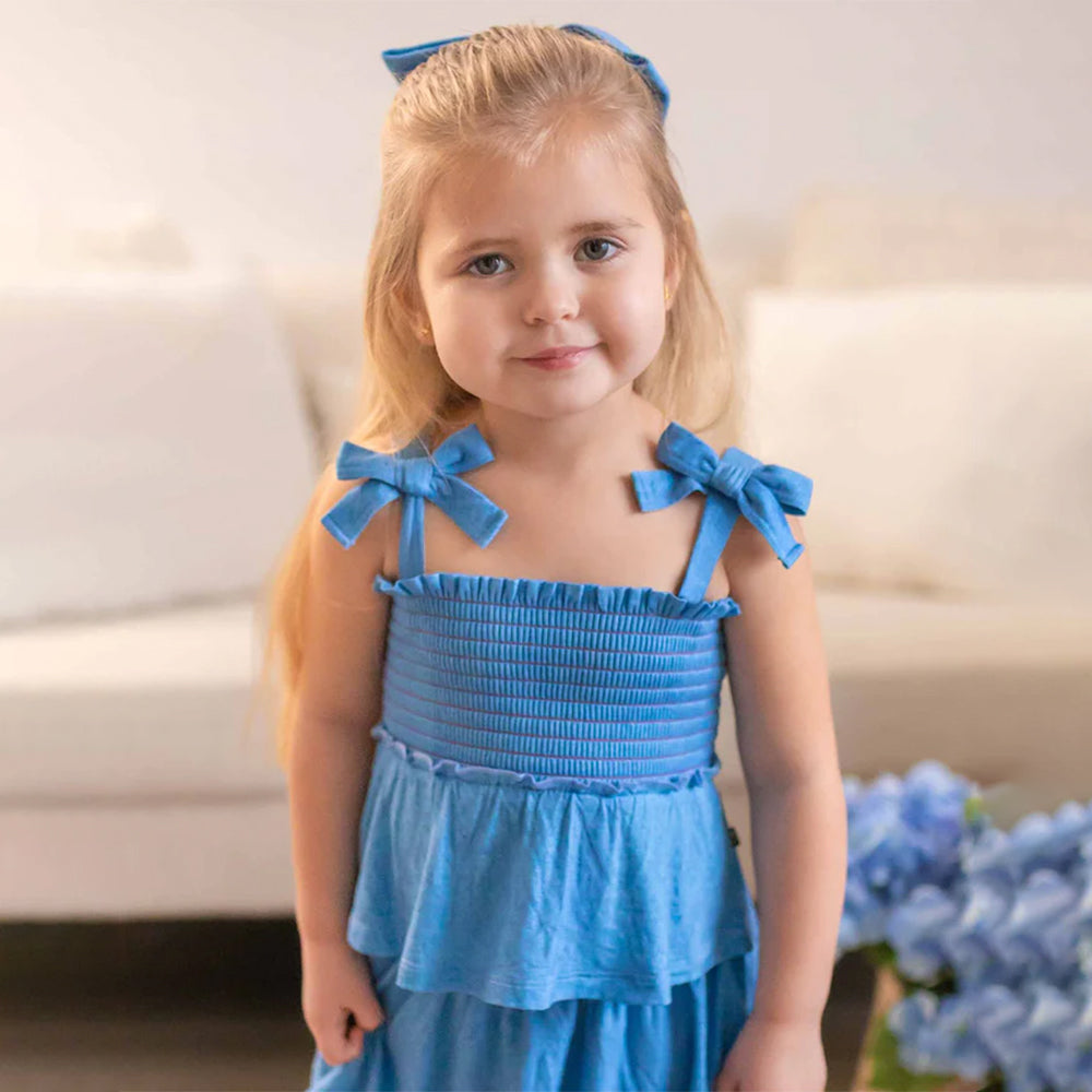 Young girl wearing a blue dress with bow straps in a softly blurred indoor setting.