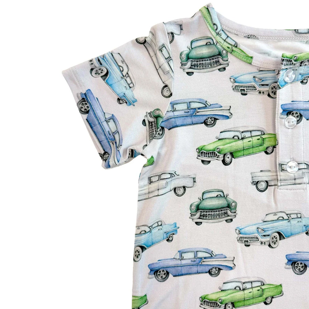 A close-up of a short-sleeve baby romper or bodysuit with an all-over vintage car print on a light cream background. The print features retro cars in light blue, medium blue, green, and white, with a buttoned chest pocket.