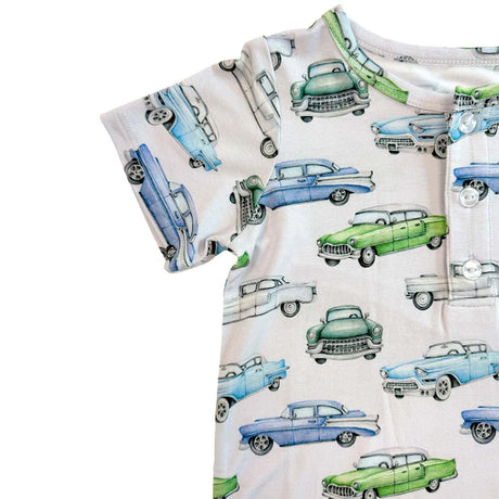 A close-up of a short-sleeve baby romper or bodysuit with an all-over vintage car print on a light cream background. The print features retro cars in light blue, medium blue, green, and white, with a buttoned chest pocket.