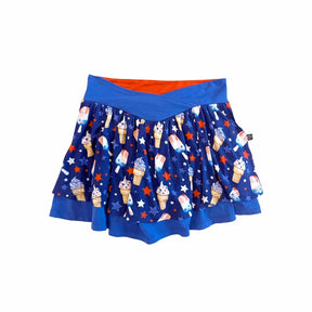 A blue pleated skater skirt featuring an all-over patriotic ice cream and star pattern, with a solid blue waistband and a bright orange inner lining visible at the top, shown against a plain white background.