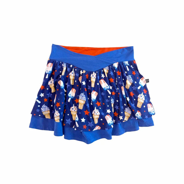 A blue pleated skater skirt featuring an all-over patriotic ice cream and star pattern, with a solid blue waistband and a bright orange inner lining visible at the top, shown against a plain white background.