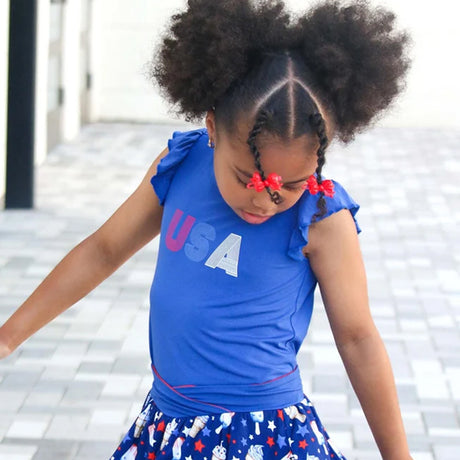 A young girl with high-pitched hair and braids wearing a blue ruffled short-sleeve top with 'USA' printed on the chest, paired with a blue patterned skirt, captured in an outdoor setting.