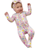 A smiling baby lies on a woven bassinet cushioned with white muslin blankets, wearing a pink footed one-piece jumpsuit with a pastel floral and paisley print, plus a matching bow headband.