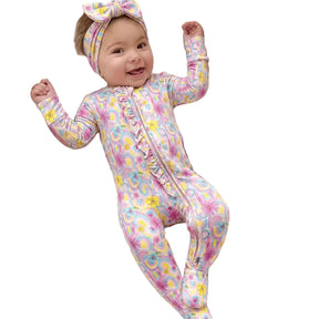 A smiling baby lies on a woven bassinet cushioned with white muslin blankets, wearing a pink footed one-piece jumpsuit with a pastel floral and paisley print, plus a matching bow headband.