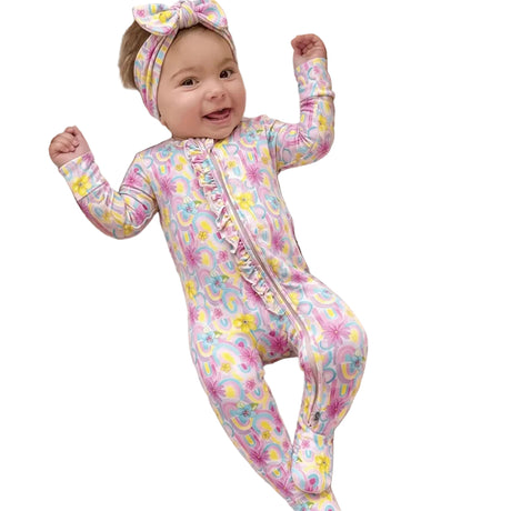 A smiling baby lies on a woven bassinet cushioned with white muslin blankets, wearing a pink footed one-piece jumpsuit with a pastel floral and paisley print, plus a matching bow headband.