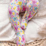 A pair of pastel rainbow and floral patterned footed slippers with a zipper closure, displayed on a textured white blanket.