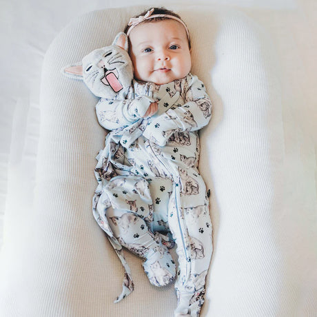Baby wrapped in a patterned swaddle with a soft toy, lying on a white cushion.