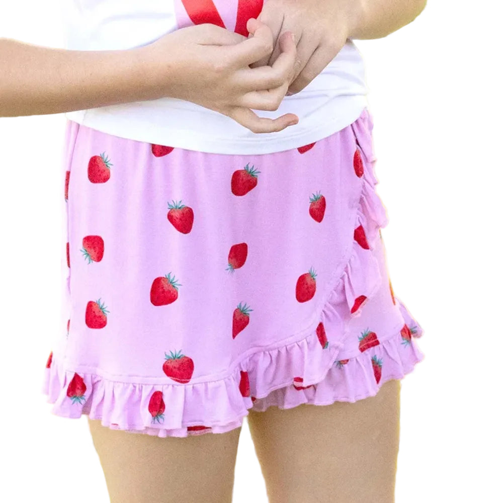 A child wearing a light pink ruffled skirt with a red strawberry print, paired with a white top, standing outdoors against a blurred green grass background.