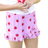 A child wearing a light pink ruffled skirt with a red strawberry print, paired with a white top, standing outdoors against a blurred green grass background.