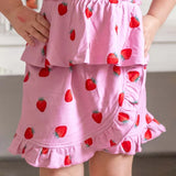 A close-up of a child wearing a pink tiered skirt with a strawberry print, showing the ruffled hem and side pockets.