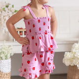 A young girl wearing a pink ruffled sleeveless dress with a strawberry print, standing in a softly decorated indoor setting.