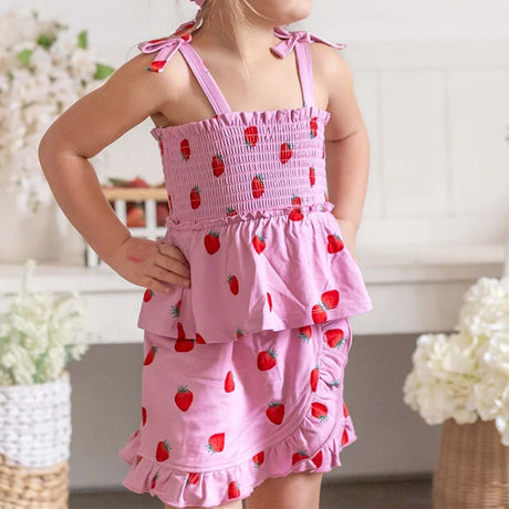 A young girl wearing a pink ruffled sleeveless dress with a strawberry print, standing in a softly decorated indoor setting.