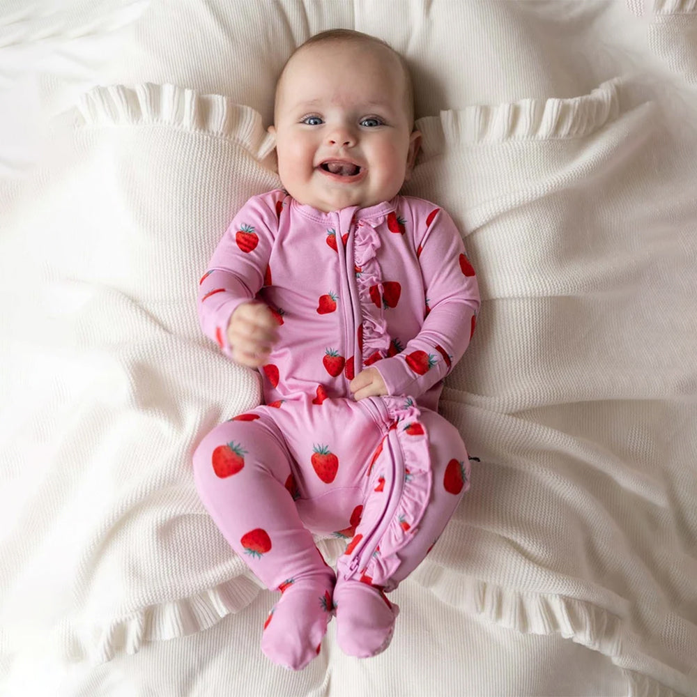 A baby lying on a cream-colored textured blanket, wearing a pink footed sleeper with a red strawberry print.