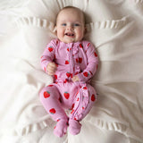 A baby lying on a cream-colored textured blanket, wearing a pink footed sleeper with a red strawberry print.