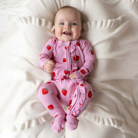 A baby lying on a cream-colored textured blanket, wearing a pink footed sleeper with a red strawberry print.