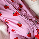 A close-up of a pink baby garment, likely a footmuff or sleeper, featuring a red strawberry print and ruffled trim, laid on a soft white textured blanket.