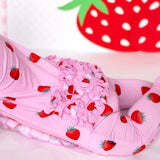 Pink children's leggings with a strawberry print and a ruffled waistband, shown on a child lying on a white surface with a large strawberry decoration in the background.