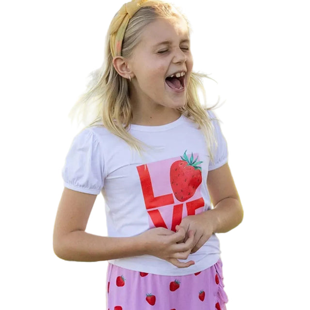 A young girl laughing outdoors on a grassy lawn near water, wearing a white short-sleeve T-shirt with a large pink and red strawberry graphic and the word LOVE, paired with a pink skirt with strawberry prints.