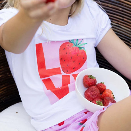 A child wearing a white graphic T-shirt with a large red strawberry and pink 'LOVE' lettering, holding a white bowl of fresh strawberries while sitting outdoors.