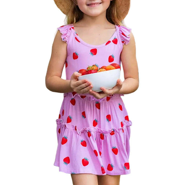 A young girl wearing a pink sleeveless dress with a red strawberry print, holding a white bowl of fresh strawberries in an outdoor garden setting.