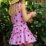 A young girl wearing a pink sleeveless dress with a red strawberry print, viewed from behind as she holds a small bowl of strawberries outdoors in front of green foliage.