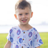 A young boy smiling outdoors, wearing a light blue short-sleeve henley shirt with a baseball-themed print featuring blue and red baseballs, bats, and white uniforms.