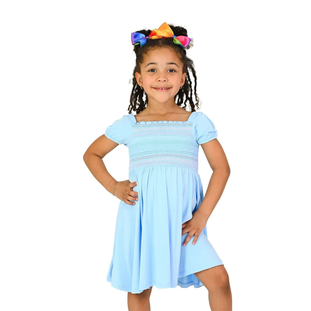 A young girl with braided hair wearing a light blue smocked dress and a colorful rainbow bow headband, posing against a solid white background.