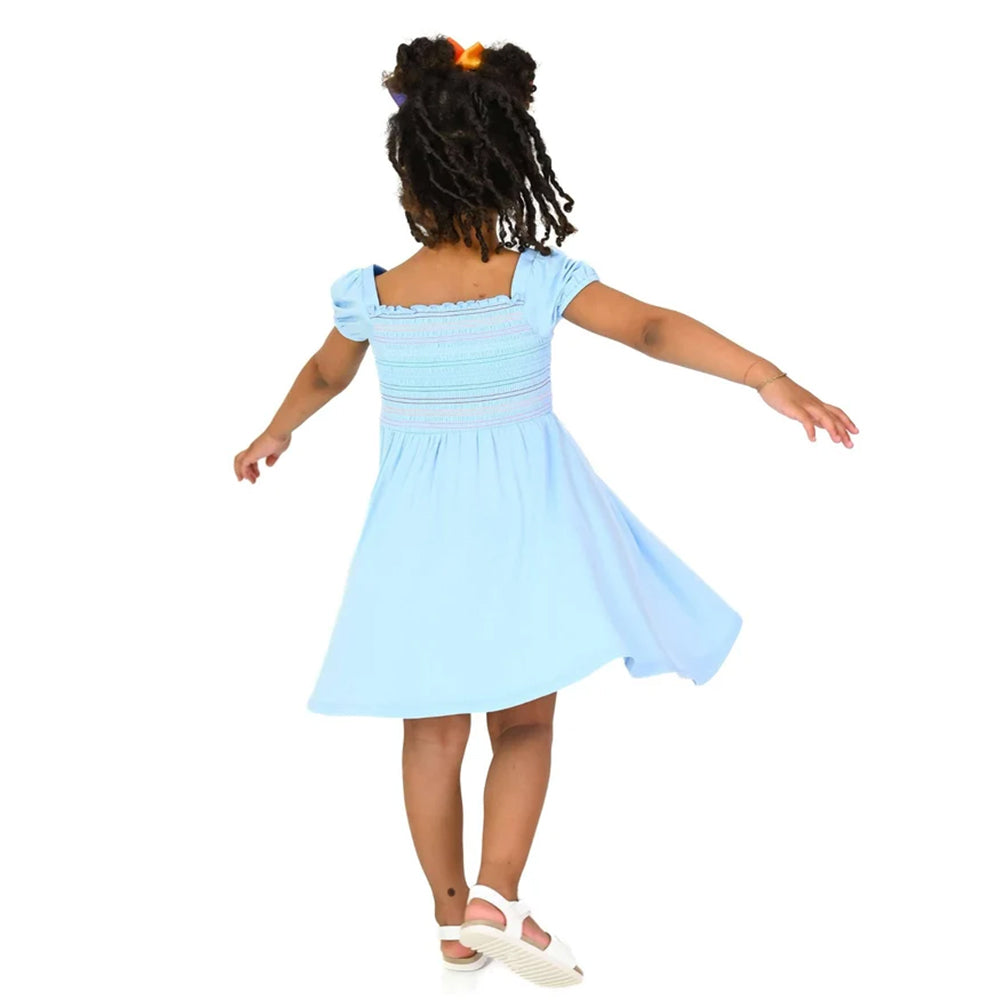 A young girl with dark curly hair is shown from behind wearing a light blue smocked dress with short puff sleeves and white wedge sandals, set against a solid white background.